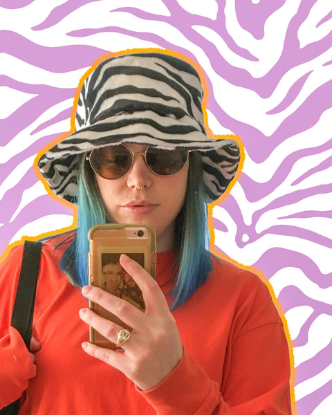 Young woman wears the Zebra print faux fur bucket hat. She is taking a selfie in the mirror.