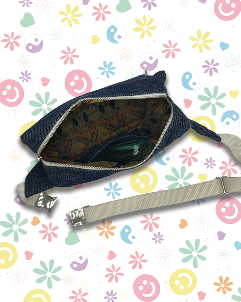 A dark blue denim bumbag with an open zipper, showing a Liberty fabric lining and a coin pocket, with a light beige adjustable strap. 