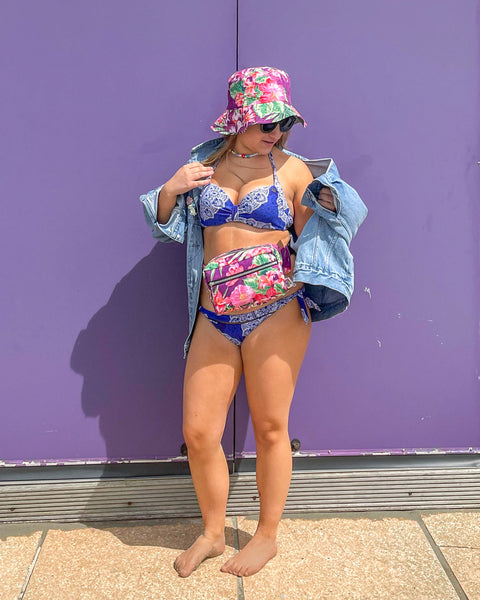 Young woman wearing the purple & pink tropical floral reversible bucket hat and matching tropical bumbag. She is standing in front of a beach hut. 
