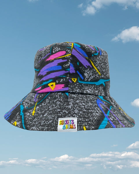 Product shot - black retro 80s bucket hat with streaks of purple, blue and pink all over showing Buckets & Bums woven label