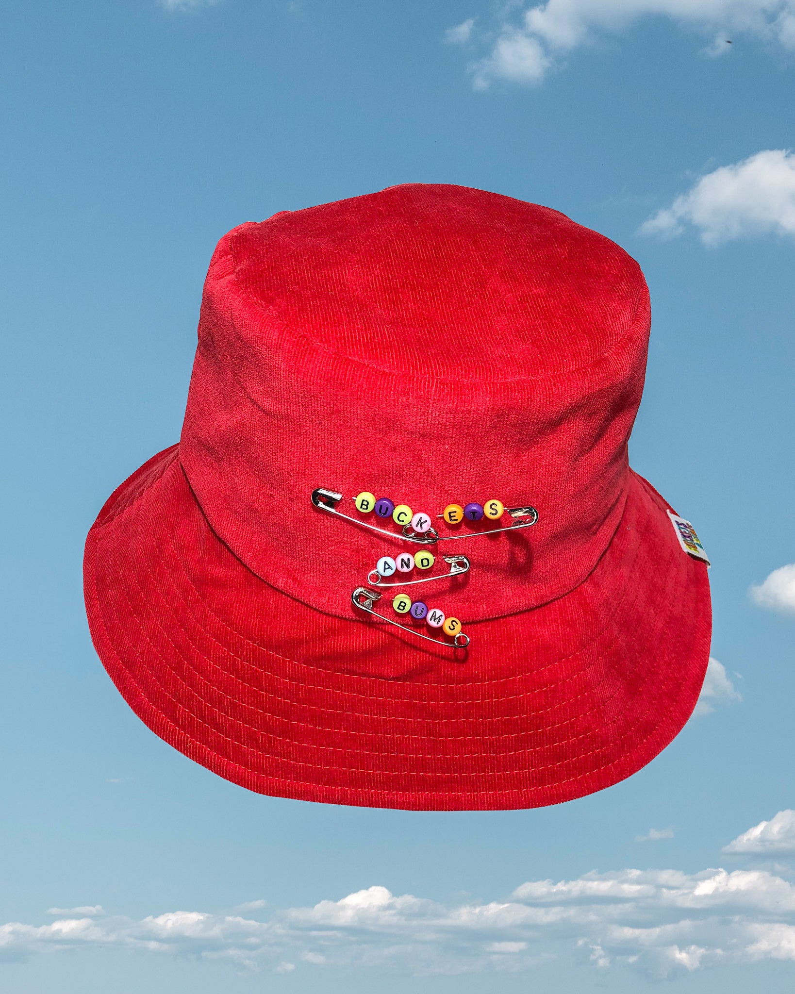 Red Safety Pin Corduroy Bucket Hat. Safety pins have colourful letter beads on which spell out Buckets & Bums