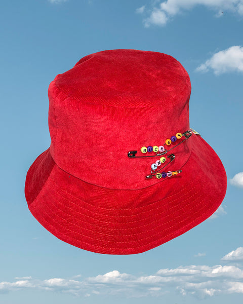 Red Safety Pin Corduroy Bucket Hat. Safety pins have colourful letter beads on which spell out Buckets & Bums